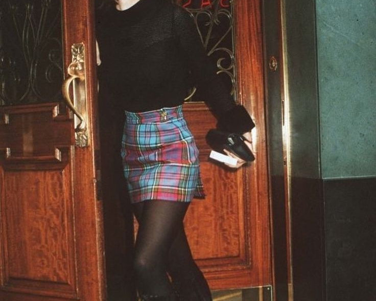 A person wearing a black long-sleeve top, a colorful plaid mini skirt, black tights, and black lace-up knee-high boots, standing in a doorway holding a small black bag.