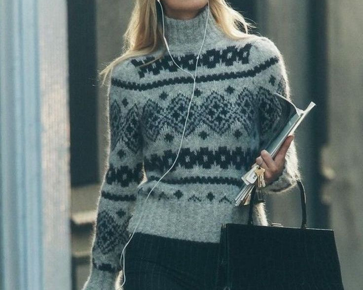 A woman wearing a gray and black patterned knit turtleneck sweater, dark pinstriped trousers, and carrying a black tote bag while walking outdoors.