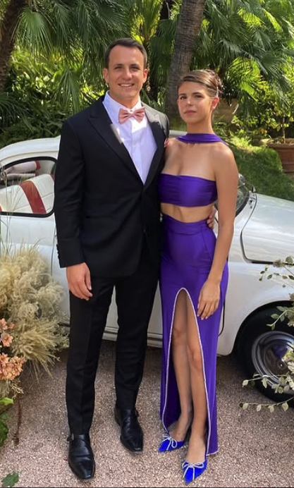 Man in a suit and woman in a purple dress standing in front of a vintage car with greenery.